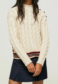 Strickpullover in Creme mit Kabelmuster, geripptem Saum mit rot-weißen Streifen, Rundhals und Knopfdetails an den Schultern. Figurbetonte Passform.