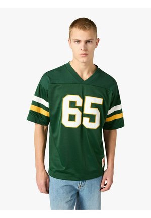 Giovane uomo con capelli biondi corti che indossa una maglia da calcio verde con il numero 65, strisce bianche e gialle sulle maniche e jeans azzurri.