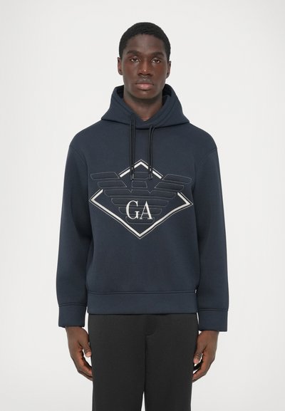 Hombre vistiendo una sudadera con capucha de un azul marino oscuro con un gran logotipo de contorno blanco y las letras "GA" sobre un fondo liso claro.