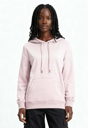 Femme aux longs cheveux tressés portant un sweat à capuche rose clair et un pantalon cargo noir, debout devant un fond blanc uni.