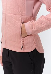 Svetle růžová bunda s texturovaným geometrickým vzorem, s kapsami na zip a elastickými manžetami, spárovaná s černými kalhotami s detaily na zip.