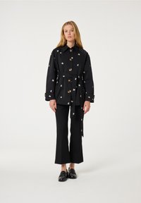 Zwart ceinture jasje met bloemenborduursel, knopen aan de voorkant en lange mouwen, gecombineerd met zwarte strakke broek en zwarte instappers.