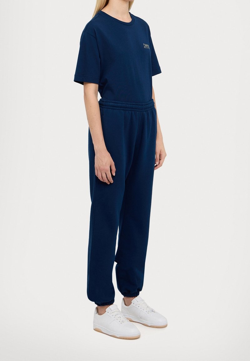 Navy blauwe katoenen t-shirt en bijpassende joggers met elastische tailleband en boorden. Witte sneakers met lichtbruine zolen. Eenvoudig, casual ontwerp.