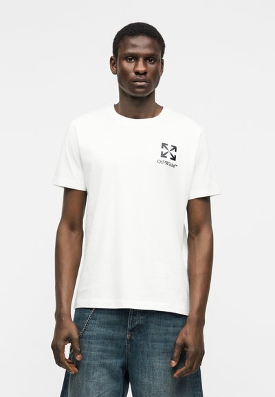 Άνδρας που φοράει λευκό t-shirt Off-White με μαύρο λογότυπο στο στήθος και ανοιχτό μπλε τζιν, στέκεται μπροστά σε απλό φωτεινό φόντο.