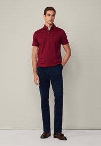 Burgundské polo tričko s límcem, které je spárováno s námořnickými kalhotami a hnědými botami. Model stojí s jednou rukou v kapse proti neutrálnímu pozadí.