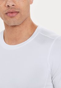 Valkoinen urheilupaita, jossa on pyöreä pääntie, lyhyet hihat ja hienovarainen teksti-brändäys olkapäällä. Materiaali vaikuttaa sileältä ja istuvalta.