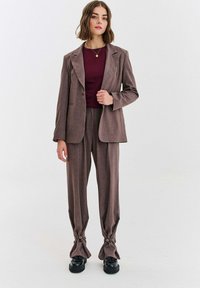 Hnedý oblekový set pozostávajúci z blejzra s výstrihom na klapky a na mieru šitých nohavíc s uzlovými manžetami a gombíkmi. Nosené cez burgundské tričko.