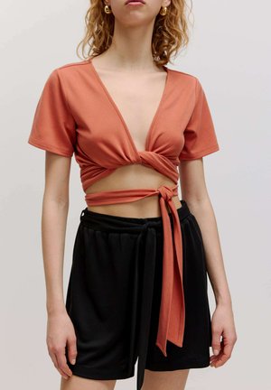 Roodbruine cropped top met een diepe V-hals en een gedraaide voorkant, gecombineerd met zwarte shorts met trekkoord. Soepele, stretchy stof.
