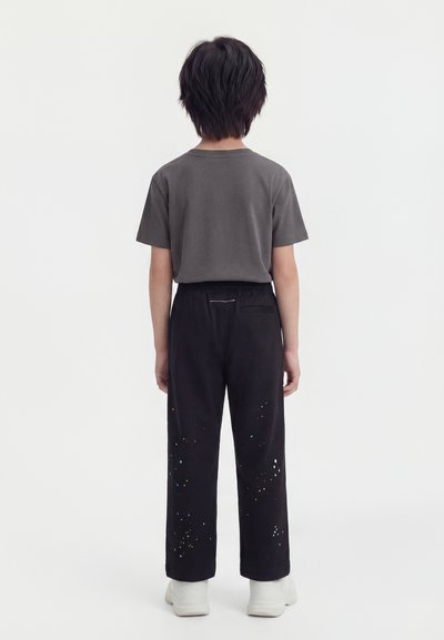 Παιδί στέκεται με την πλάτη, φοράει σκούρο γκρι μπλουζάκι, μαύρο παντελόνι με πολύχρωμες πιτσιλιές μπογιάς και λευκά παπούτσια.