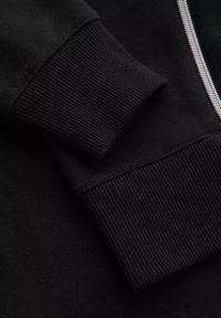 Gros plan d'une manche de sweat-shirt noire avec un poignet côtelé et une fermeture éclair métallique visible sur le corps du vêtement.