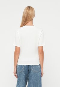 T-shirt blanc à manches courtes avec un col rond, en tissu de coton doux. Vu de dos, associé à un pantalon en denim bleu à motif.