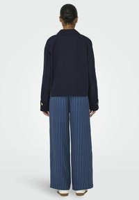 Blazer navy con una vestibilità leggermente ampia, abbinato a pantaloni a gamba larga a righe blu e bianche. Presenta dettagli in bottone oro sulle maniche.