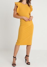 Gul figursydd klänning med volangärmar, knälång, tillverkad av elastisk tyg. Matchad med svarta högklackade sandaler. Slät yta.