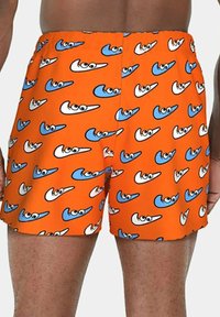 Pantaloni corti arancioni brillanti con un motivo ripetitivo di forme a swoosh blu e bianche con occhi di cartone, indossati da una persona con pelle scura.