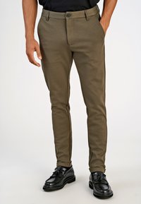 Khakihosen mit schmaler Passform, aus weichem Stoff, mit Knopfschluss, seitlichen Taschen und schwarzen seitlichen Akzenten. Getragen mit schwarzen Schuhen.