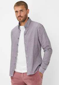 Camicia a maniche lunghe con un motivo a quadri rosso e blu, con colletto. Indossata sopra una t-shirt bianca, abbinata a pantaloni rossi chiari.