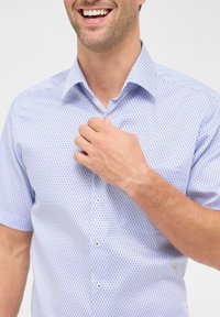Kurzärmliges Hemd aus hellblauem Stoff mit einem kleinen geometrischen Muster, ausgestattet mit einem spitzen Kragen und einer Knopfleiste vorne.