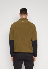 Fleece pullover i olivengrøn med sorte nylon ærmer, høj krave og et lille logo på ryggen. Blød tekstur og tætsiddende design.