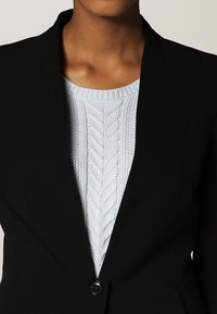 Svart blazermed djup halsringning och en enkel knappstängning. Under finns en ljusblå stickad tröja med kabelmönster synlig.