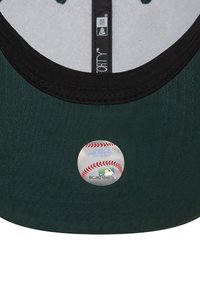 Zöld baseball sapka fekete napellenzővel, szövet textúrával, és egy kör alakú holografikus matricával, amelyen baseball dizájn látható az alsó oldalán.