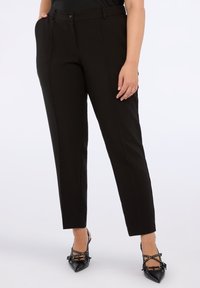 Kvinna klädd i skräddarsydda svarta byxor och svarta högklackade skor med spetsiga tår och nitar som korsar sig, stående mot en vit bakgrund.