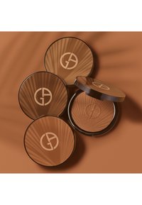 Quatre compacts de bronzer circulaires de différentes nuances de brun avec un motif texturé, chacun arborant un logo sur le couvercle, posés sur un fond chaud.