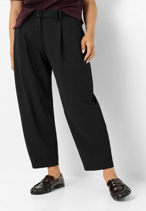 Person trägt schwarze, weite, knöchellange Hosen, dunkle Loafers und ein dunkles Oberteil und steht vor einem einfarbigen hellen Hintergrund.