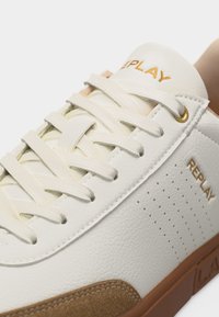 Κοντινή λήψη λευκού αθλητικού παπουτσιού Replay με λευκά κορδόνια, καφέ σκούρα τμήματα από σουέντ στη μύτη και καφέ λαστιχένια σόλα.