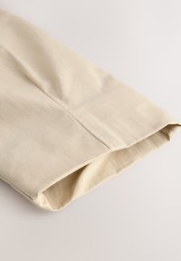 Beige katoenstof, met een gevouwen zoom die een gladde textuur en subtiele naaddetails langs de rand laat zien.