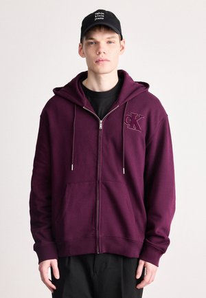 Sudadera con cremallera en color burdeos, con bolsillos frontales, cordones y logo bordado "CK". Lleva puesta sobre una camiseta negra, combinada con pantalones negros y una gorra.