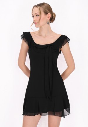 Sort kjole med flæse ved neckline, korte ærmer og en sløjfedetail. Udfoldet kant og let materiale skaber et svævende udseende.