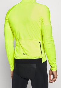 Giacca da ciclismo a maniche lunghe riflettente di colore giallo brillante, realizzata in materiale leggero e elasticizzato, con tasca posteriore con zip e logo.