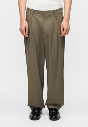 Homme portant un pantalon ample olive et des chaussures en cuir noir, se tenant devant un fond blanc uni.