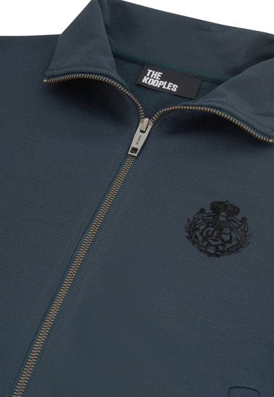 Donkerblauwe zip-up jas met een opstaande kraag, metalen rits en geborduurd embleem op de linkerkant van de borst, met het label "THE KOOPLES" aan de binnenkant van de kraag.