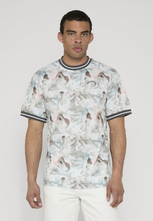 Hombre con pantalones blancos y una camisa de manga corta con estampado abstracto y cuello y puños a rayas blancas y negras, de pie quieto.