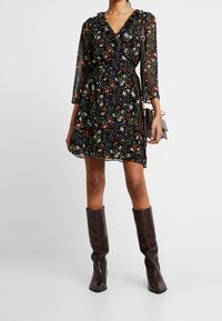 Vestido negro con flores, de manga 3/4, con escote rizado y detalle de cinturón. Combinado con botas altas hasta la rodilla, de color marrón oscuro y con estampado de cocodrilo.