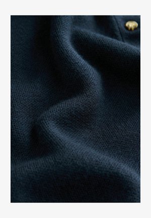 Tamsiai mėlynas megztas audinys, turintis tekstūruotą paviršių, su švelniais raukšlėmis ir matomu auksiniu mygtuko akcentu, perteikiantis švelnumą ir šilumą.