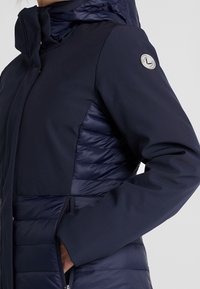 Marinblå jacka med en kombination av släta och quiltade texturer, dragkedjeförsedda fickor, hög krage och en logotypdetalj på ärmen.