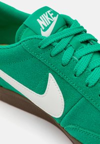 Zaļie zamša sneakeri ar teksturētu virsmu, baltu ādas swoosh un atbilstošiem šņorēm. Iezīmēts ar mīkstu mēli ar logo. Brūna gumijas zole.
