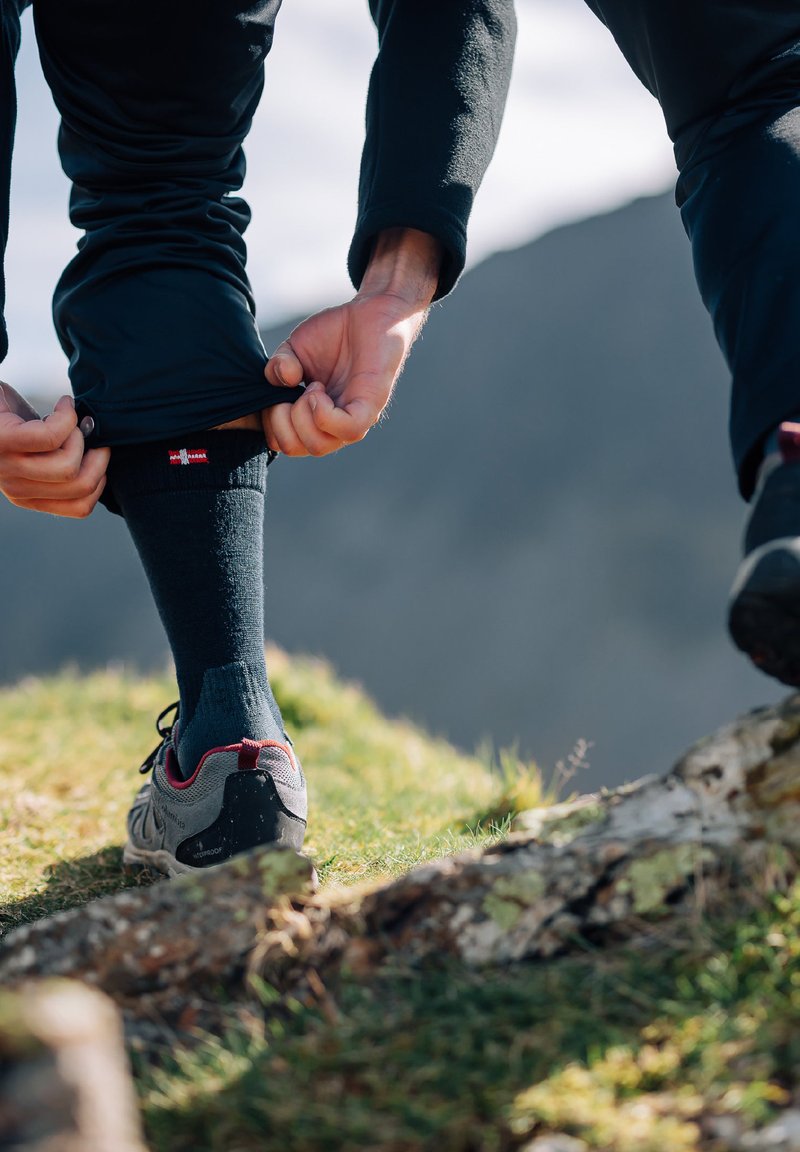 Calze invernali nere con logo, indossate con scarpe da trekking grigie con accenti rossi, su un pendio erboso. Mani che regolano i pantaloni sopra le calze.