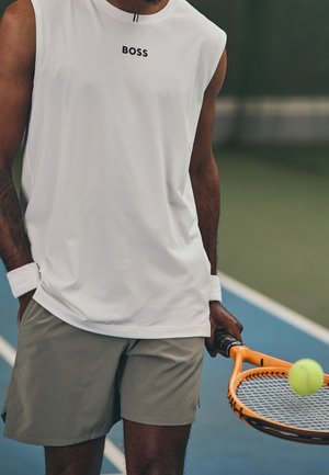 Mand iført hvid ærmeløs trøje og grå shorts, der holder en orange tennisketcher med bold på en blå tennisbane.