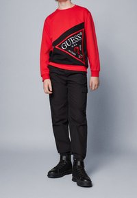 Kind in een lange rode shirt met groot zwart en rood GUESS-logo, zwarte cargobroek en zwarte high-top sneakers, staand tegen een grijze achtergrond.