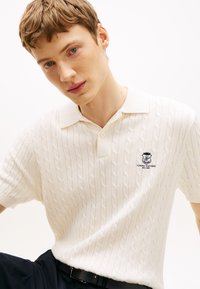 Jauns vīrietis ar īsiem matiem, tērpies baltā pinuma polo kreklā ar Tommy Hilfiger logotipu uz krūtīm, sēž pret vienkāršu fonu.