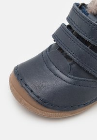 Tmavě modrá kožená bota s kulatou špičkou, dvěma suchými zipy a tanovou gumovou podrážkou. Měkké podšívka viditelná kolem okraje.