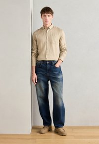 Camisa bege de mangas longas, jeans de ganga azuis com um design desbotado e sapatos beges. O modelo está posicionado contra um fundo cinza claro.