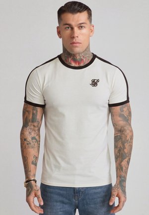 Mann mit Tätowierungen am Hals und an den Armen, trägt ein weißes, eng anliegendes T-Shirt mit schwarzem Rand und Logo, kombiniert mit blauen Jeans, steht vor einem schlichten Hintergrund.
