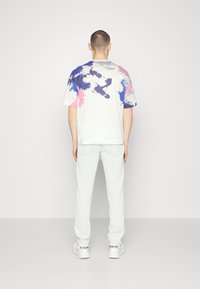 Bílé tričko s krátkým rukávem a modro-růžovými abstraktními vzory na zádech, spojené s světlými kalhotami a bílými teniskami.