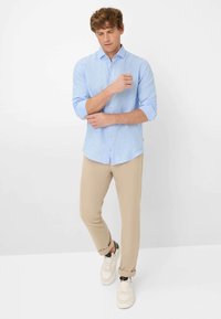 Lichtblauwe button-up shirt met opgerolde mouwen, gecombineerd met beige broek en witte schoenen. Glad materiaal en een nauwsluitende pasvorm.