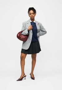 Ženska, oblečena v sivi blazer z drobnimi črtami, mornarsko modro polo majico, črno plisirano krilo in bordo petke, drži usnjeno torbico, ki se ujema z obutvijo.
