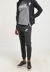 Černá zipová bunda, šedé tričko a tmavě šedé joggingové kalhoty s logy Nike. Černé tenisky s bílými podrážkami. Hladká textilie.